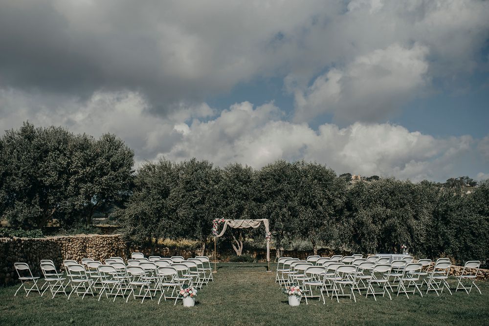 Outdoor wedding ceremony setup with white folding chairs, wooden arch, and lush olive trees under a dramatic cloudy sky.