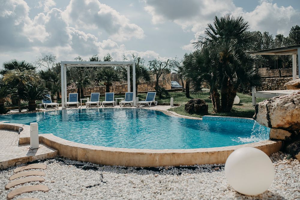 Luxury outdoor pool with lounge chairs, pergola, palm trees, and manicured gardens at a Mediterranean resort.