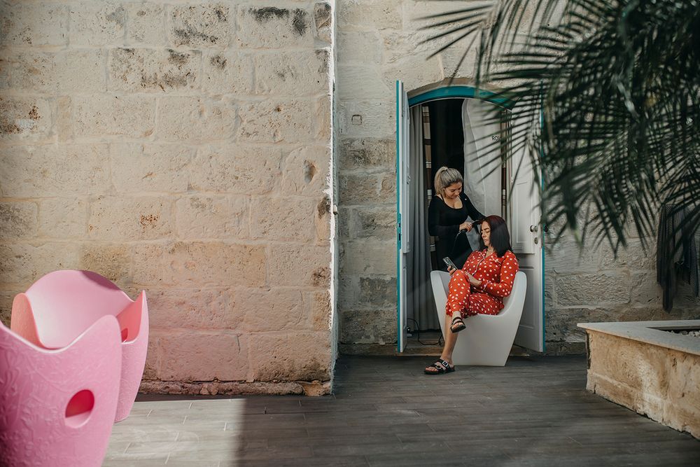 Person sitting in a white chair by a turquoise door, beside a pink decorative seat and tropical plant.