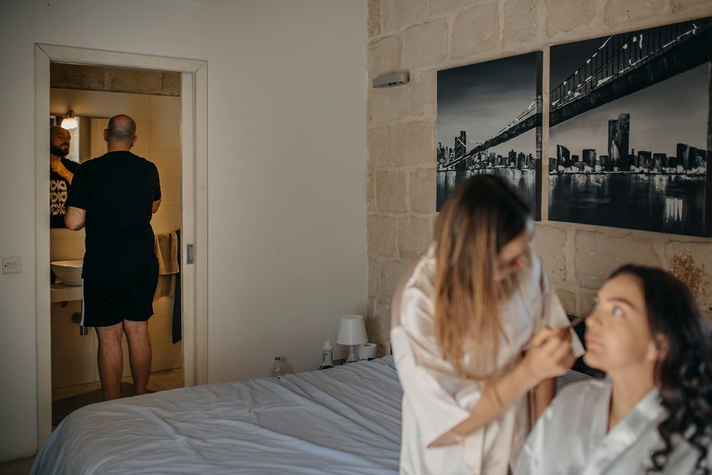 Bride getting makeup done in bedroom while man stands in doorway during wedding preparations.