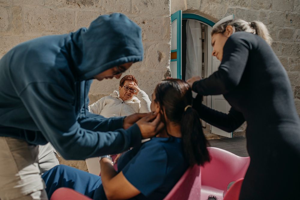 Two people styling a seated woman's hair in a salon while a man watches in the background.