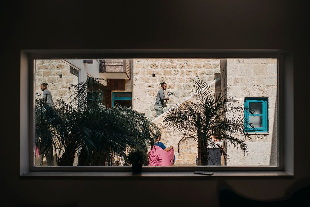 View through window of man standing near stone building with tropical plants in foreground.