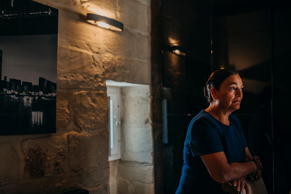 Woman in blue shirt stands thoughtfully in a dimly lit stone-walled gallery with artwork displayed on walls.