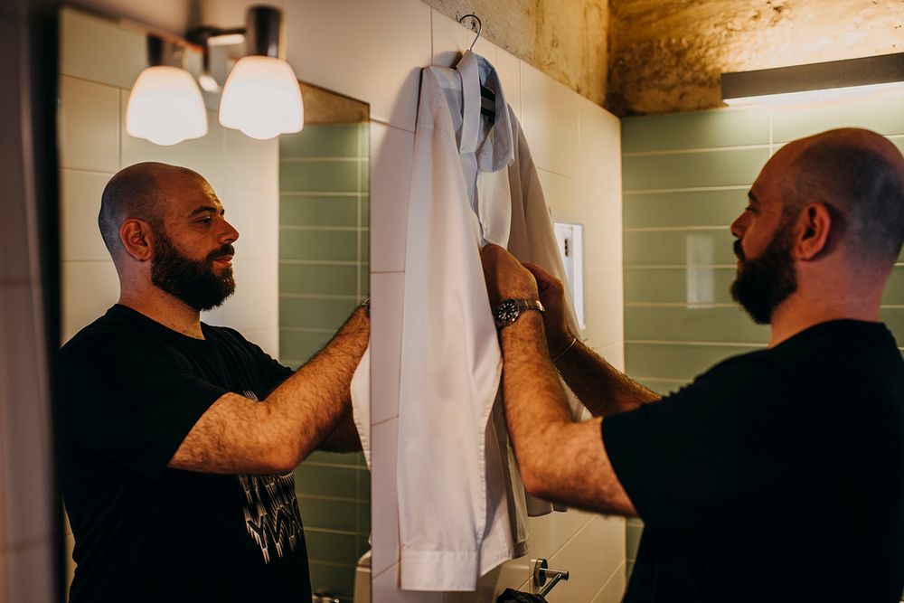 A bald man adjusts a white dress shirt on a hanger in a bathroom mirror reflection.