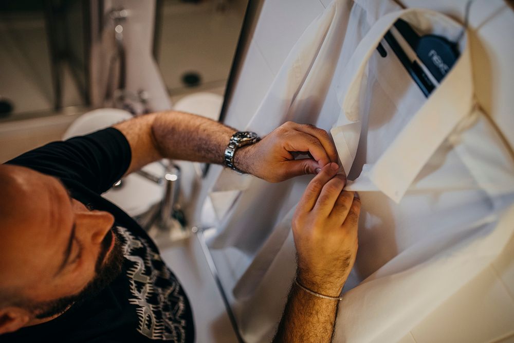 A tailor carefully adjusts a white garment on a dress form, wearing a watch while working in a professional studio.