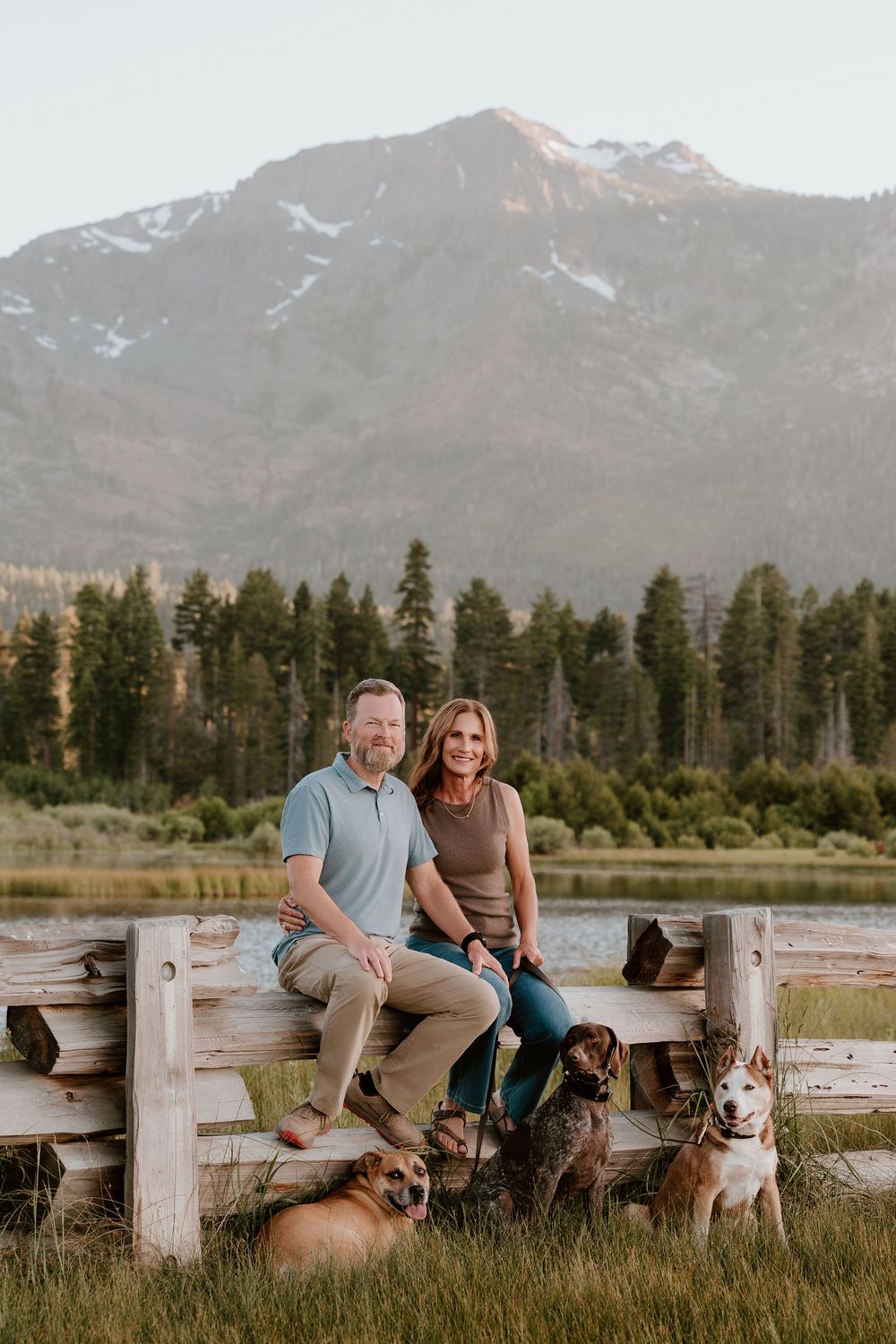 Couple sitting on rustic wooden fence with three dogs, pine trees, mountain lake, and snow-capped peak backdrop.