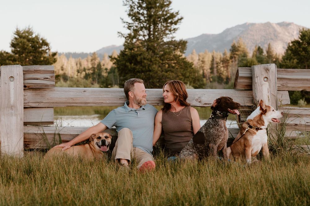 Couple sitting against rustic wooden fence in golden meadow with three dogs and mountain backdrop.