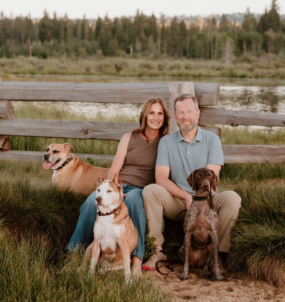 A couple sits with three dogs by a wooden fence in a scenic outdoor setting with trees and water.