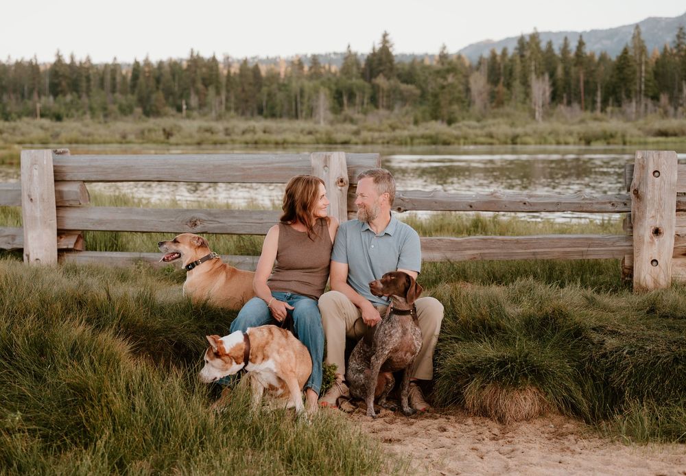Couple sitting on rustic fence with three dogs in grassy meadow with mountain lake backdrop at golden hour.
