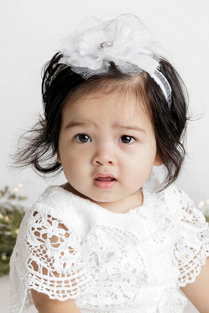 Adorable baby girl in white lace dress and floral headband with dark hair, looking curious in a studio photo.