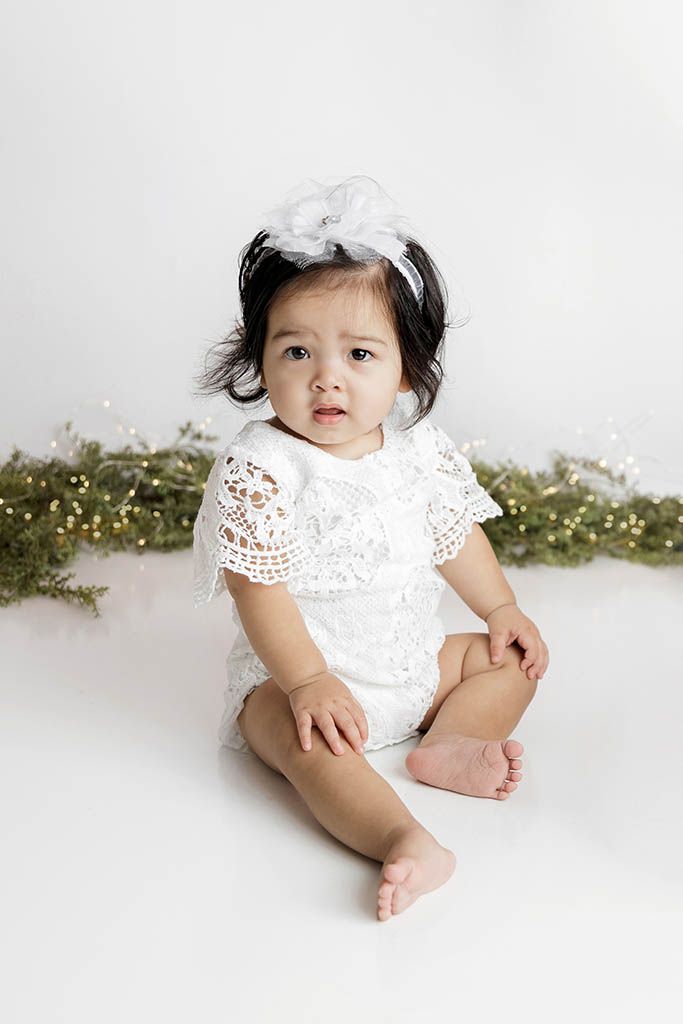 Baby girl in white lace romper and floral headband sits on white floor with greenery backdrop.