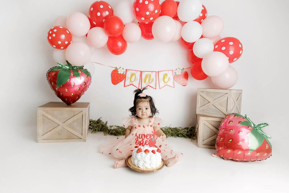 Baby girl smash cake session with strawberry theme, red and pink balloons, and wooden crates on white background.
