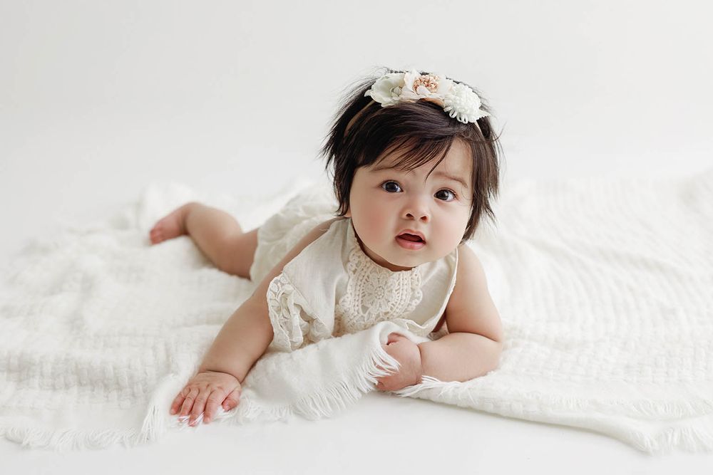 A cute baby girl in a white lace dress and floral headband lies on a soft white blanket.