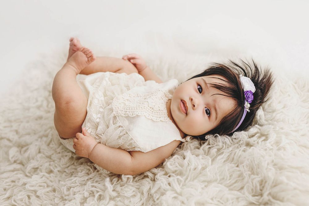 A cute baby girl in a white lace outfit lying on a fluffy cream rug with a purple floral headband.