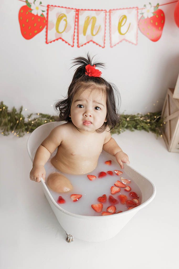 Baby girl sits in a milk bath with strawberries celebrating her first birthday with a strawberry-themed banner.