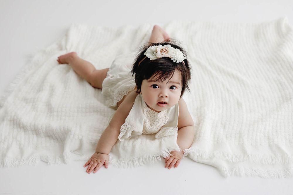 A cute baby girl in a white lace outfit and floral headband lies on a soft white blanket.