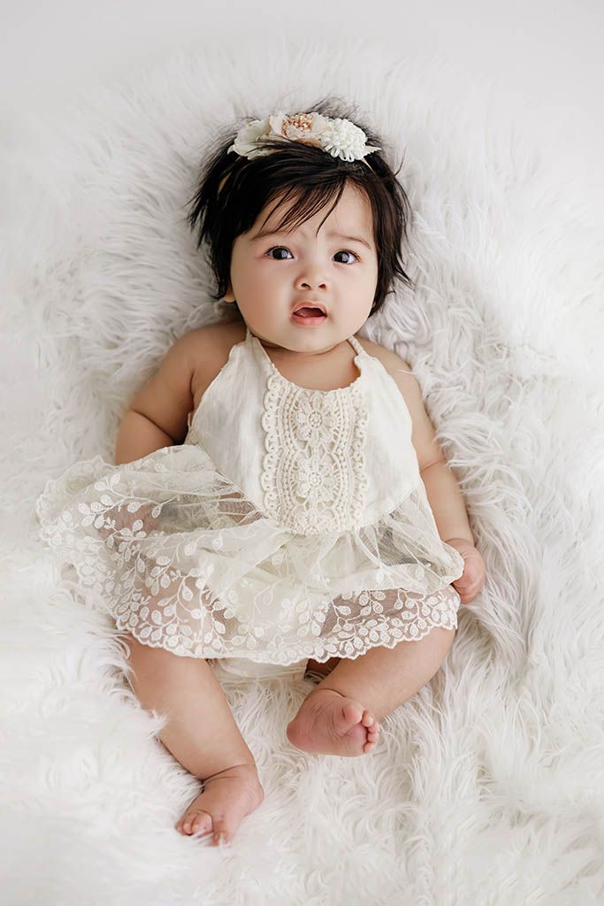 Cute baby girl wearing white lace romper and floral headband sitting on soft white fur rug.