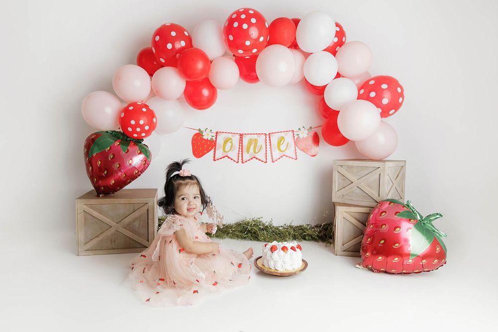 A toddler girl in a pink dress celebrates her first birthday with a strawberry cake smash setup.