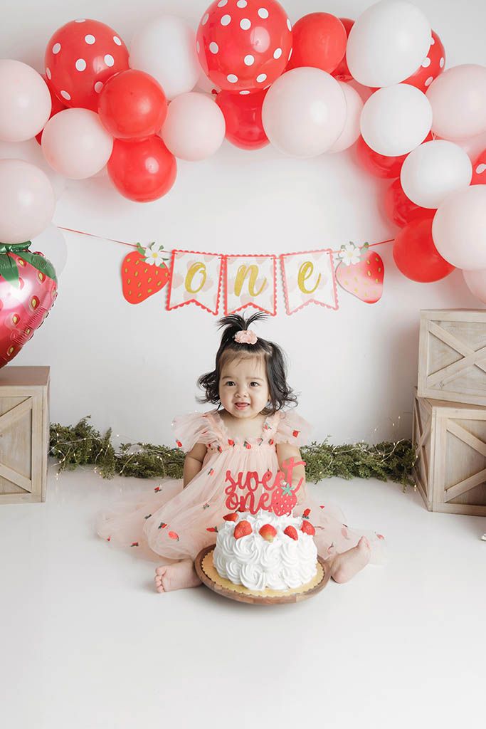 Baby girl in pink tutu sits before a strawberry-themed first birthday cake under red and white balloon arch.