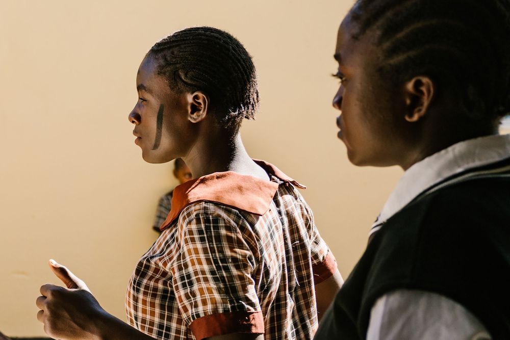 Two African schoolgirls in uniforms stand attentively in a classroom, one holding a paper.