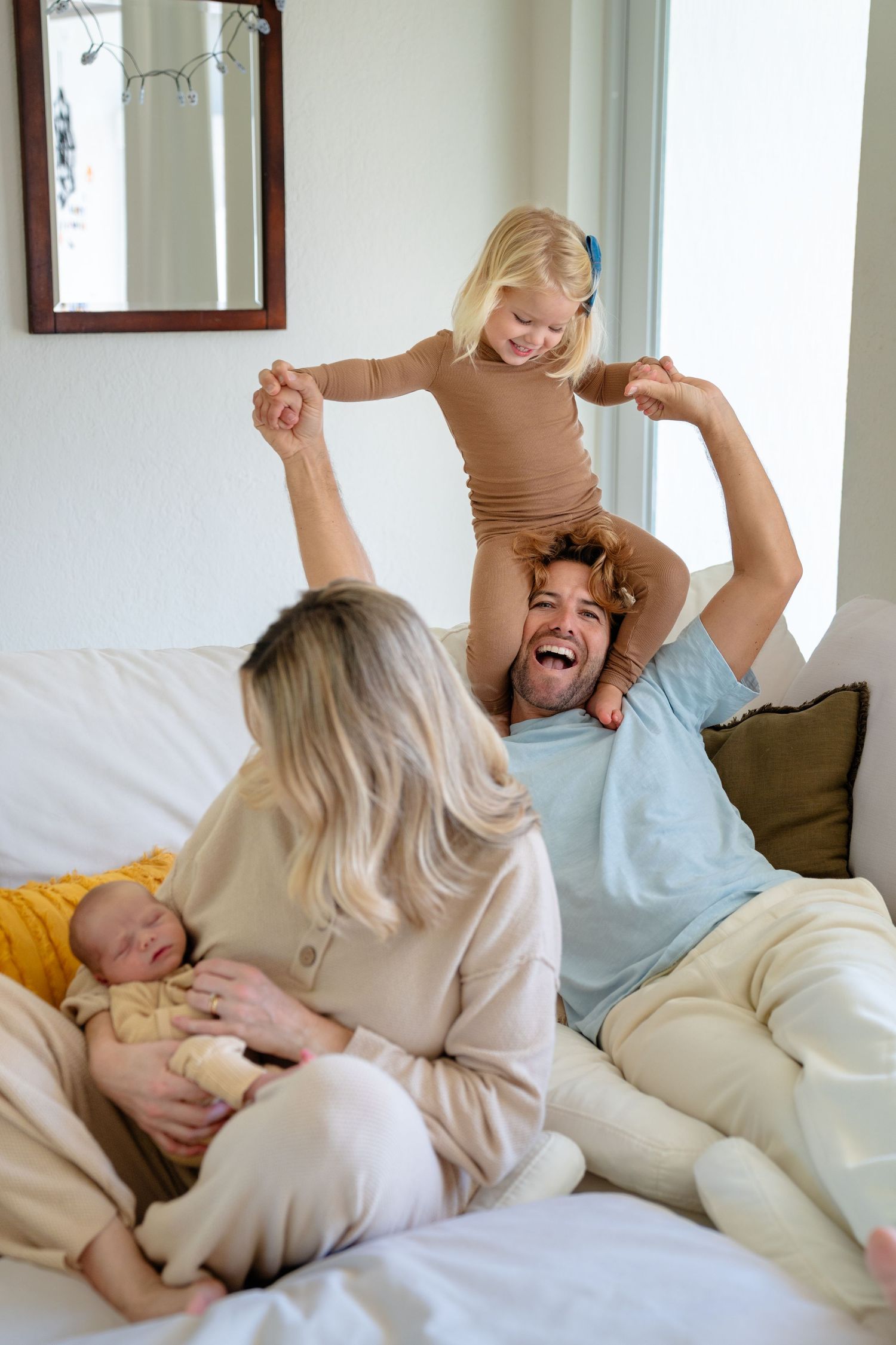 Happy family on sofa, dad laughing with toddler on shoulders, mom holding newborn baby at home.