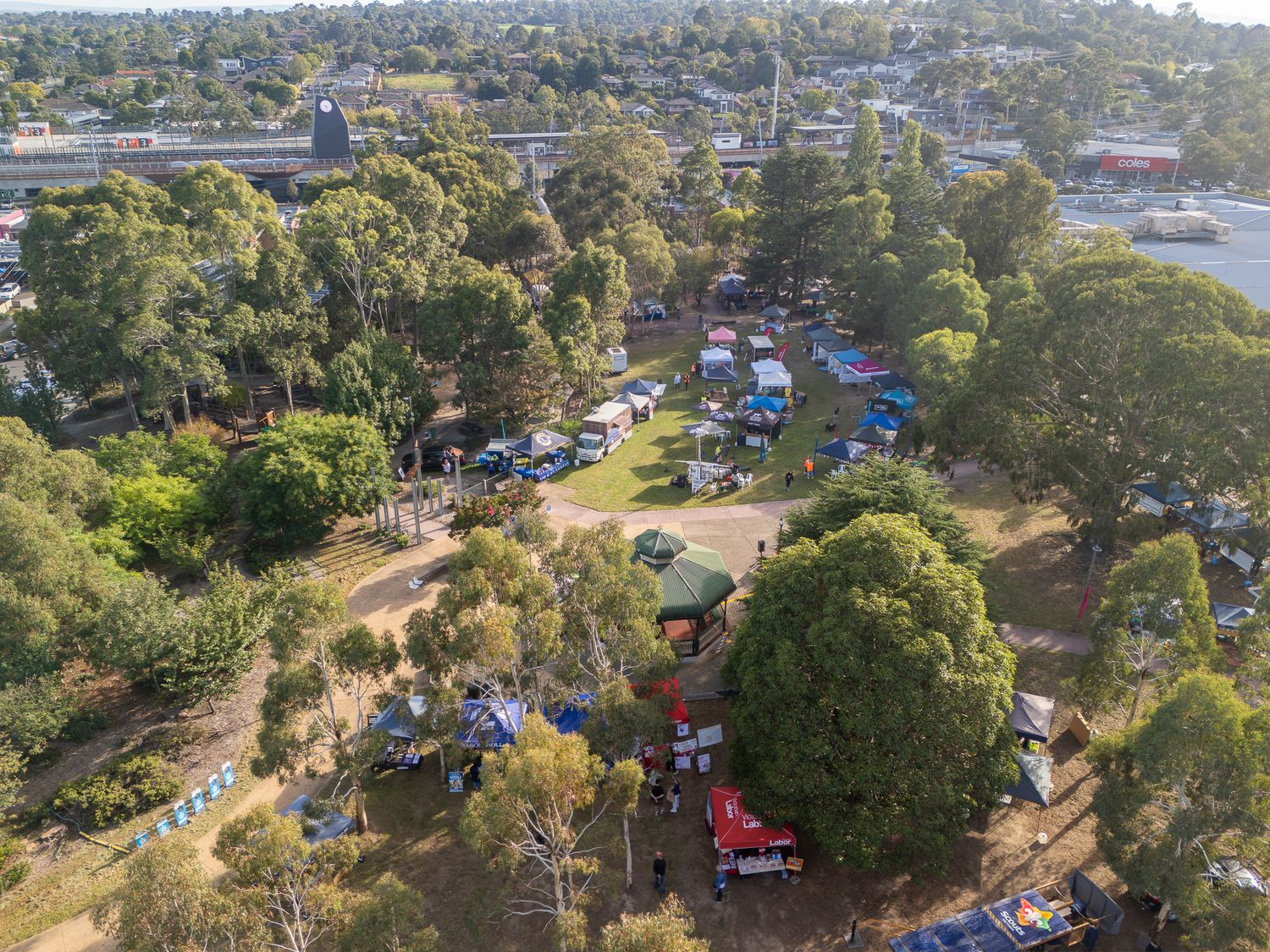 Aerial view of a lush green park with tents, vehicles, and market stalls surrounded by tall eucalyptus trees in a suburban area.