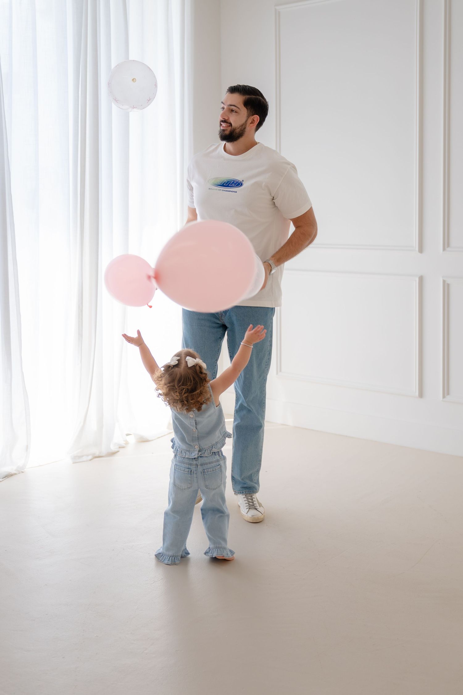 Father and daughter playing with pink and white balloons in a bright white room.