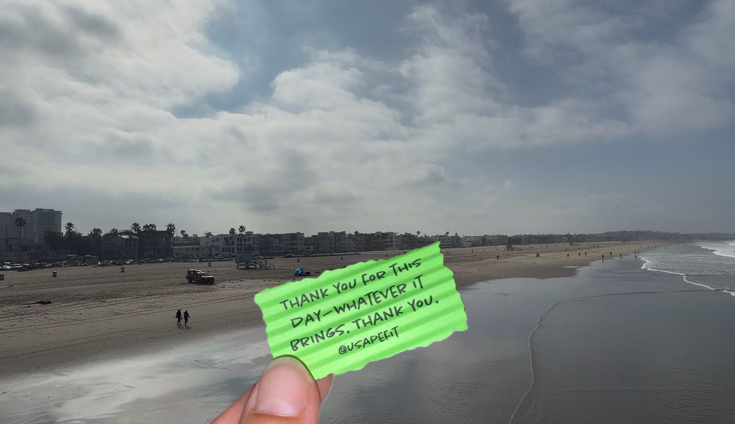 Hand holding a green note card on a sandy beach with ocean waves and cloudy sky in the background.