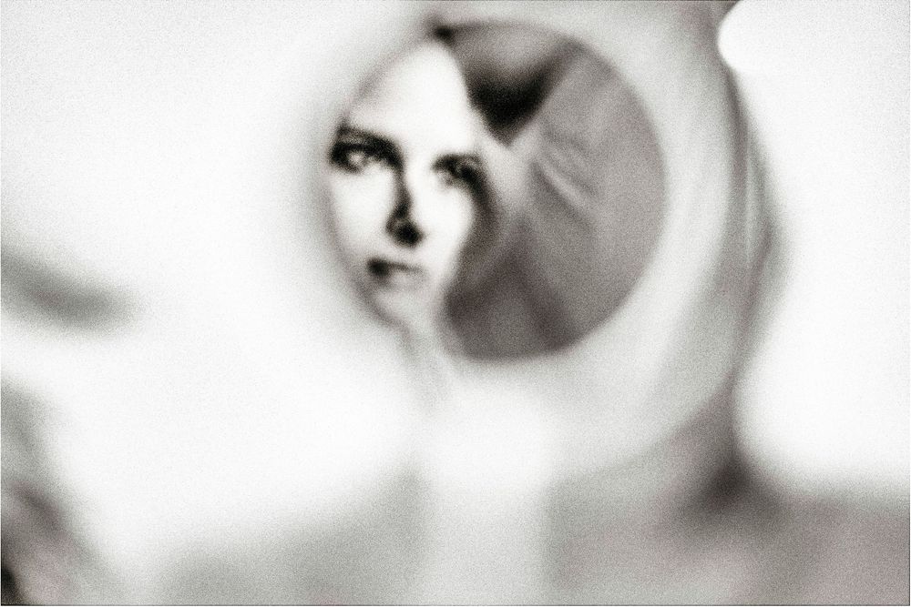 Black and white photo of a woman's face reflected in a small round mirror, artistic blurred bokeh effect.