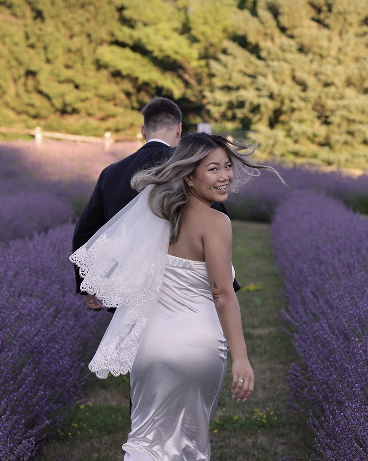 Smiling bride in white gown with veil walking through purple lavender field with groom.