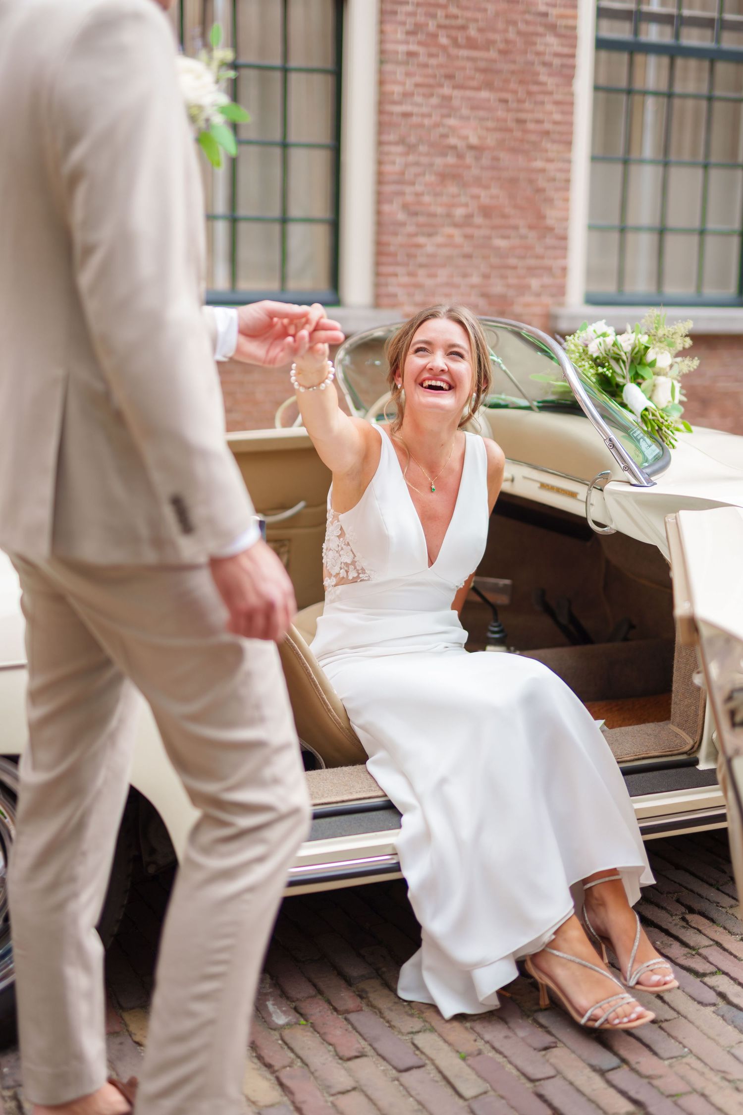 Laughing bride in white dress sits on vintage car door, reaching up to groom in beige suit on cobblestone street.
