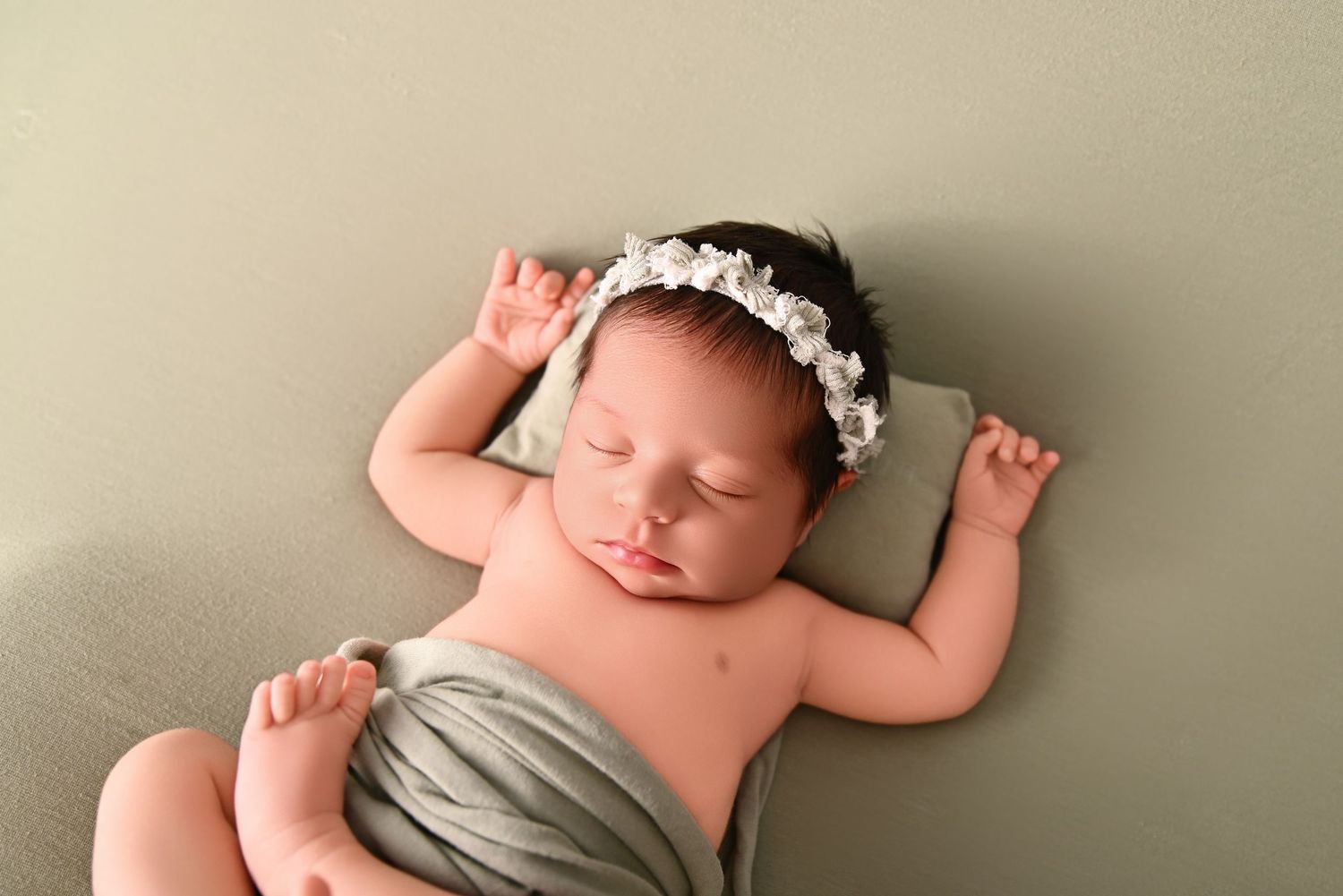 Sleeping newborn baby girl wearing a white floral headband, wrapped in sage green fabric on a matching green backdrop.