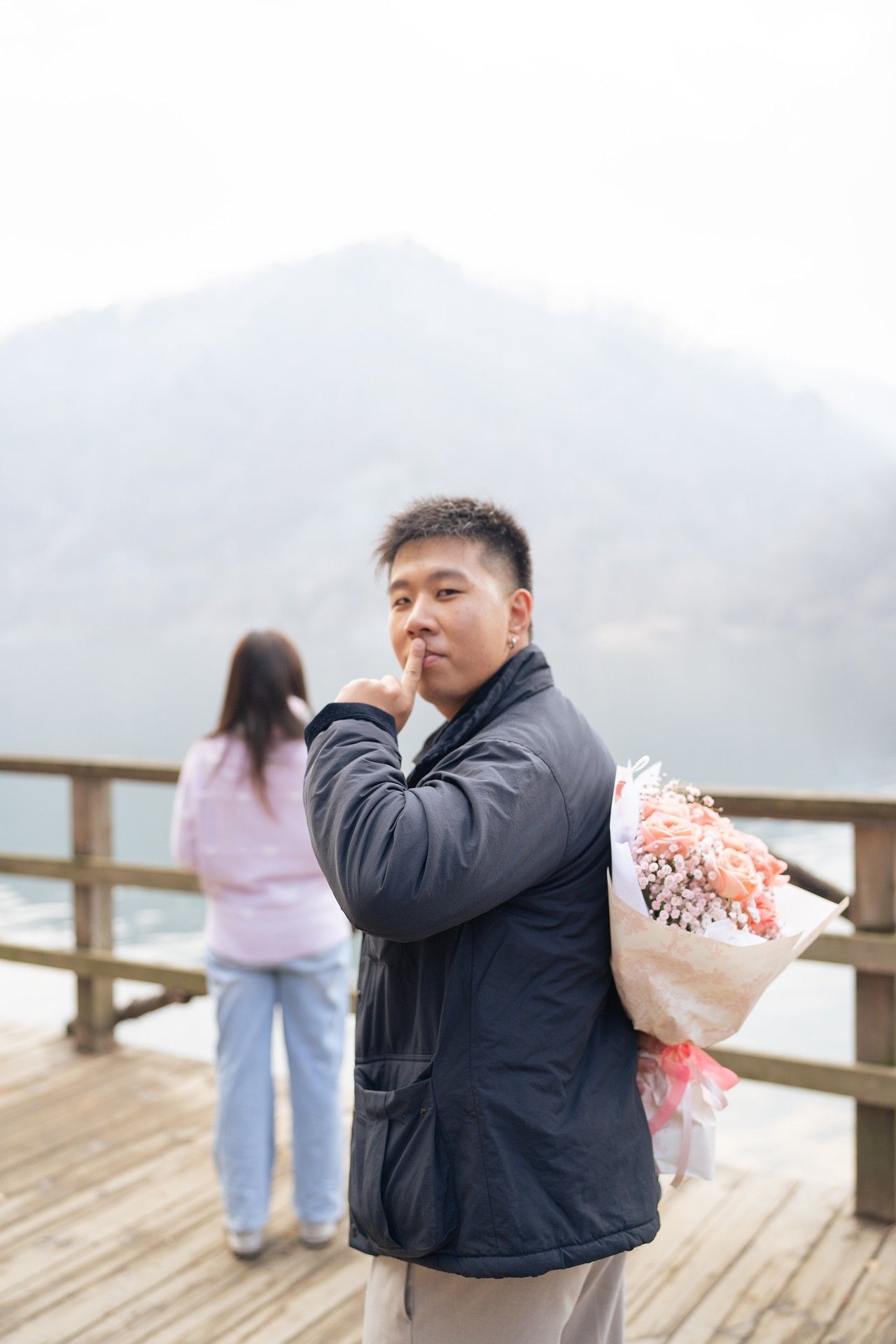 Man holding flower bouquet on wooden deck with mountain lake backdrop, appearing to plan a romantic surprise proposal.
