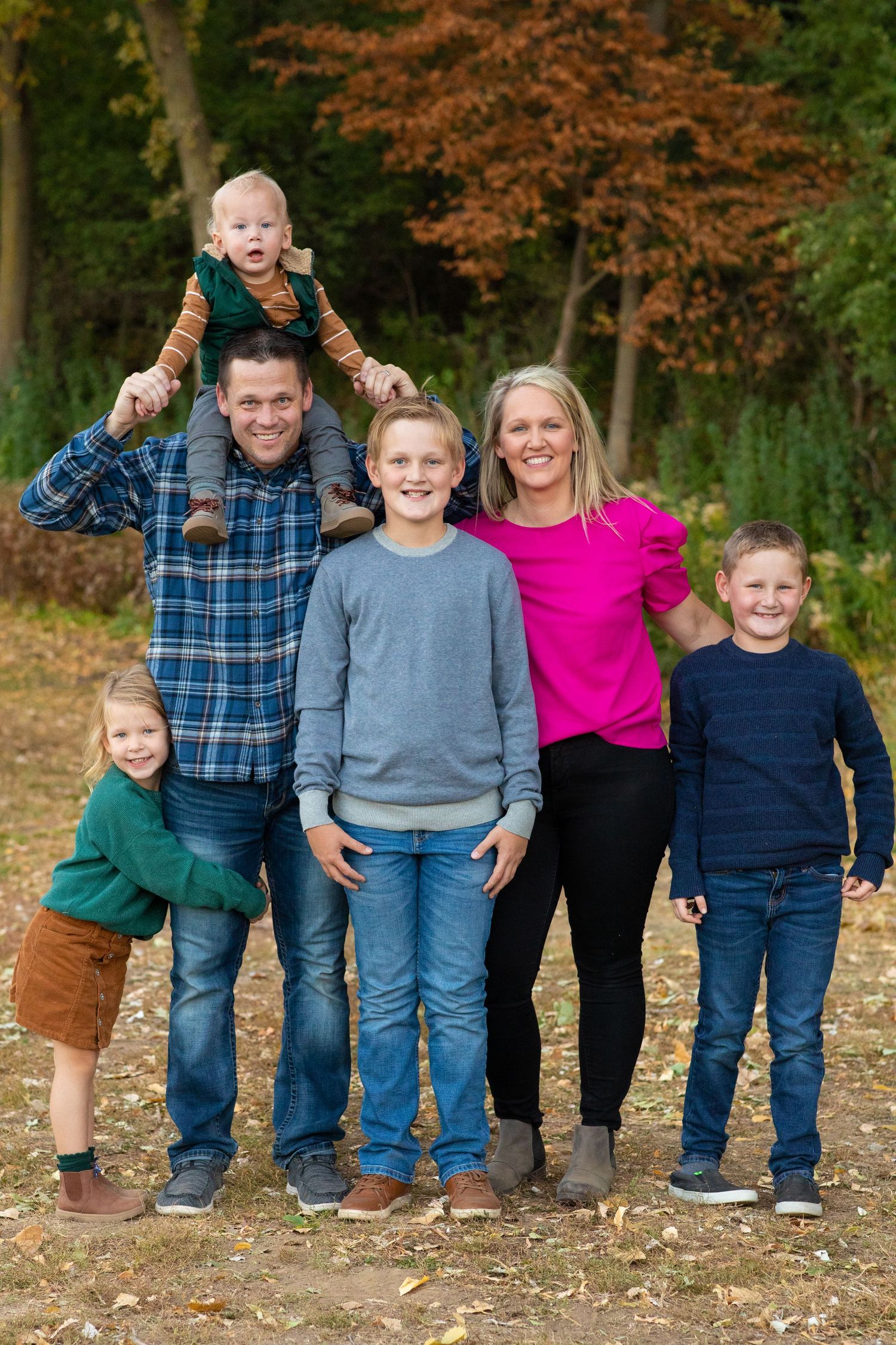Family photo shoot with everyone wearing casual clothing posing together on fallen autumn leaves in an outdoor setting.