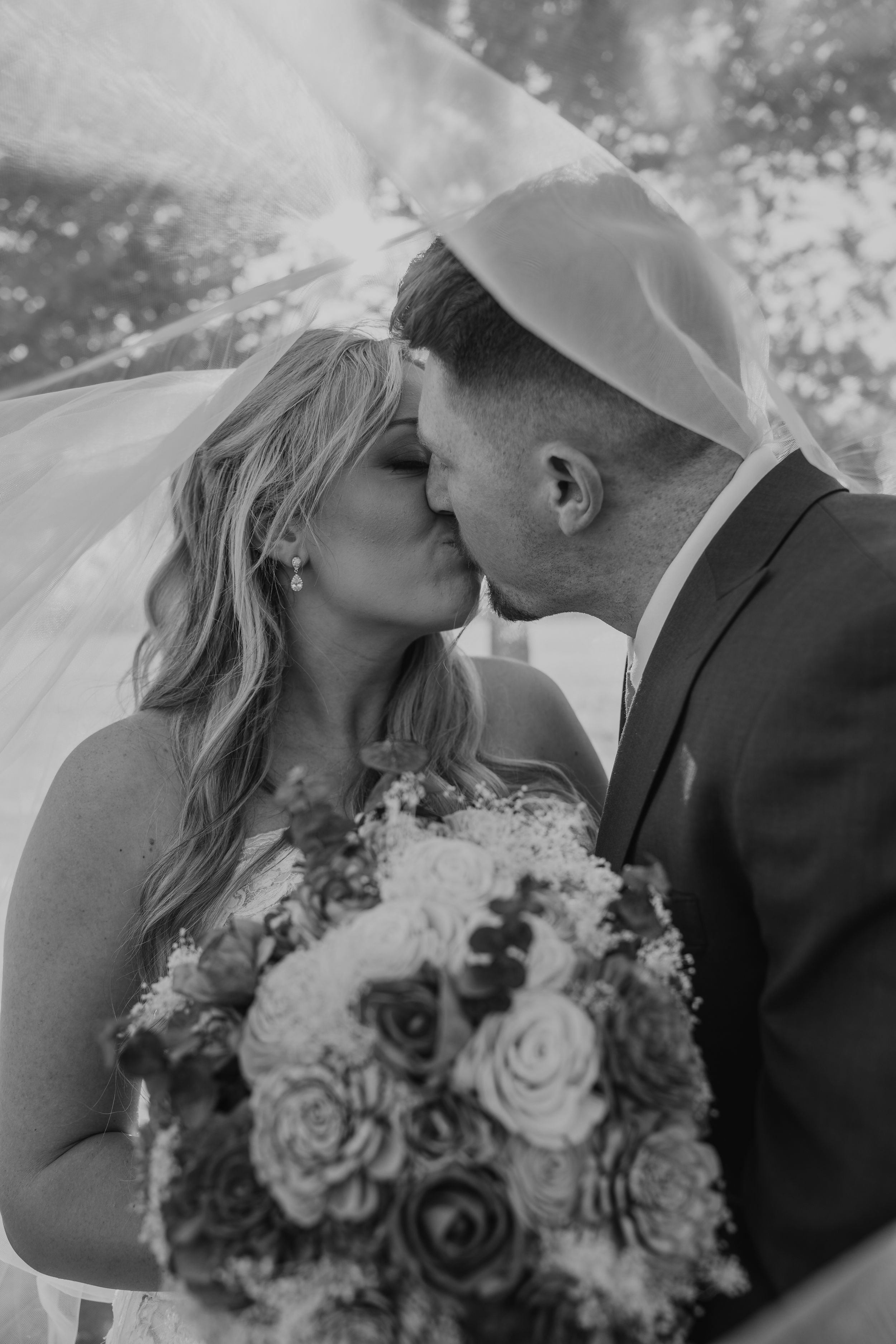 Bride and groom share a romantic kiss under a wedding veil in a dreamy black and white portrait.