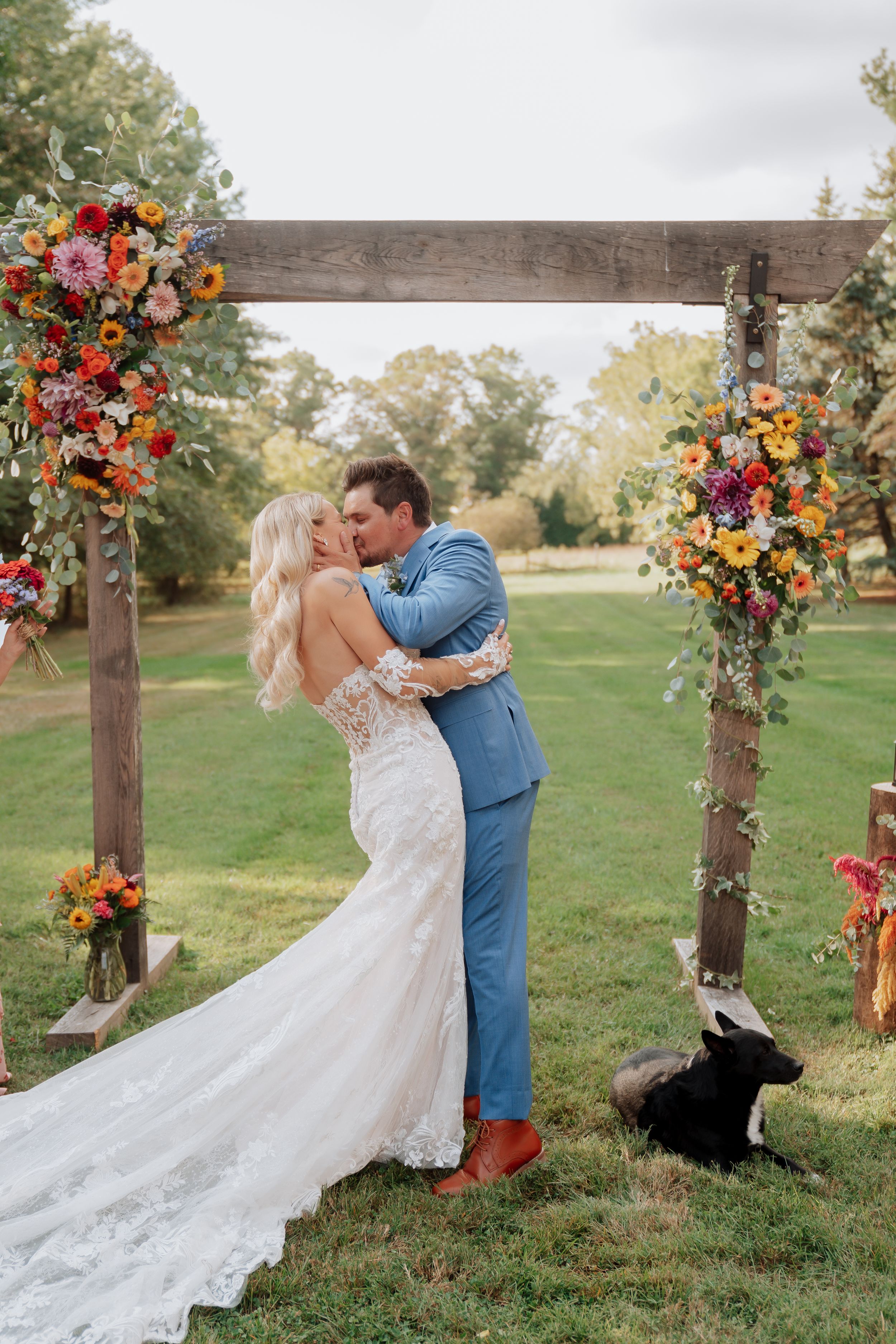 Bride and groom share first kiss under rustic wooden arch adorned with vibrant flowers at outdoor wedding.