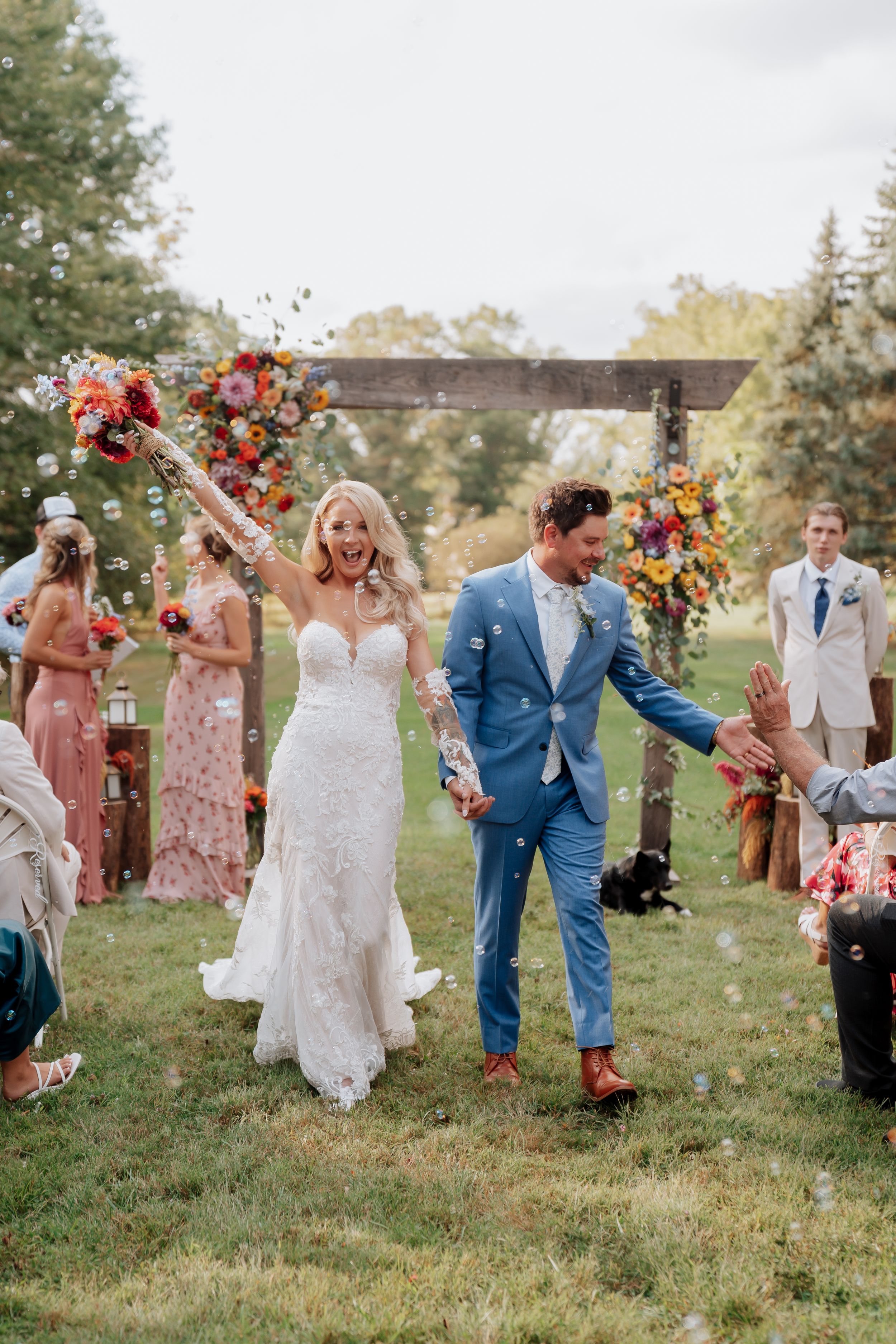 Joyful bride and groom walk back down aisle as guests blow bubbles at colorful outdoor wedding ceremony.