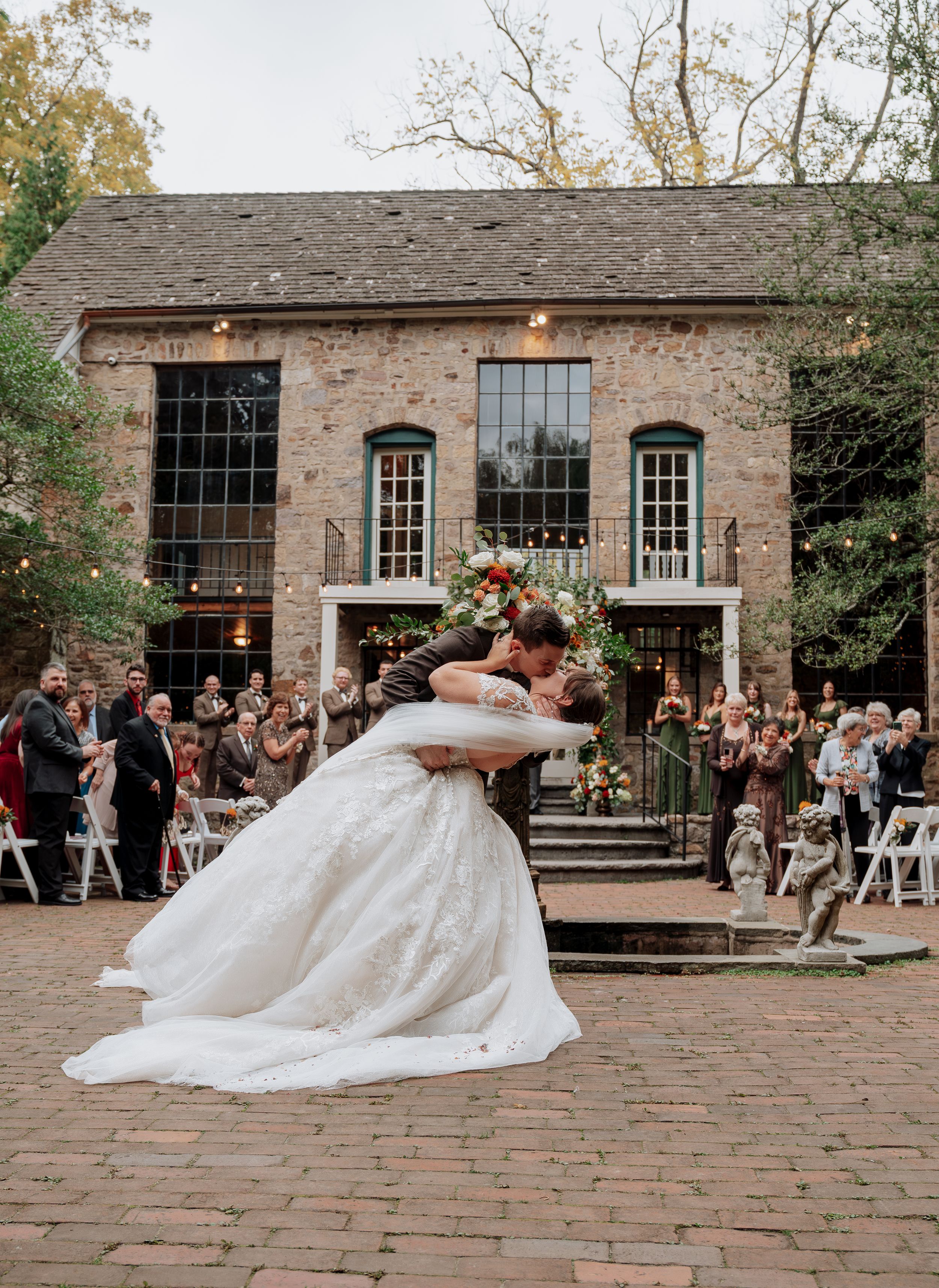 Groom dips bride in romantic kiss on brick courtyard with guests and string lights behind them.