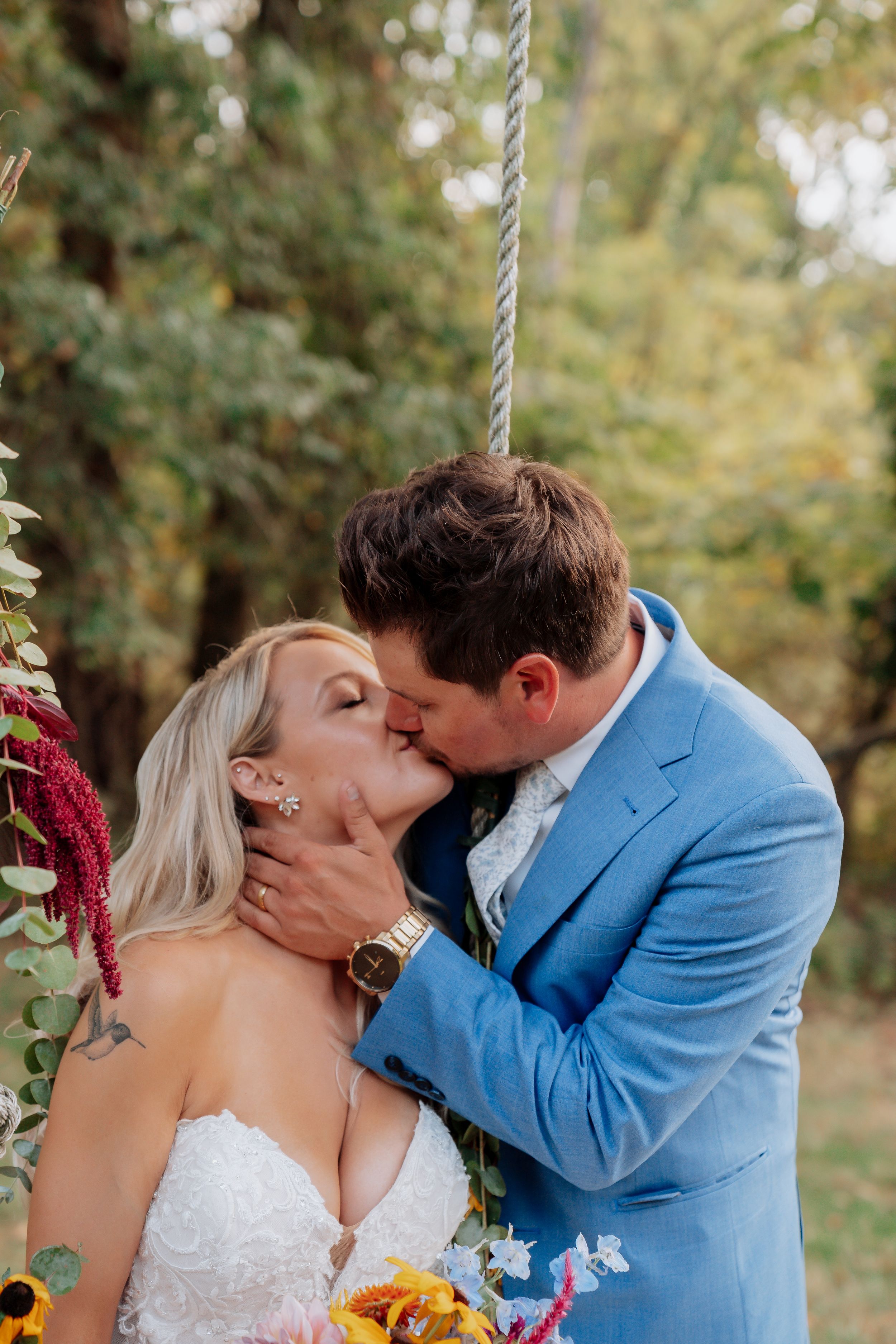 Bride and groom share passionate kiss on wooden swing surrounded by sunflowers and greenery at fall wedding.