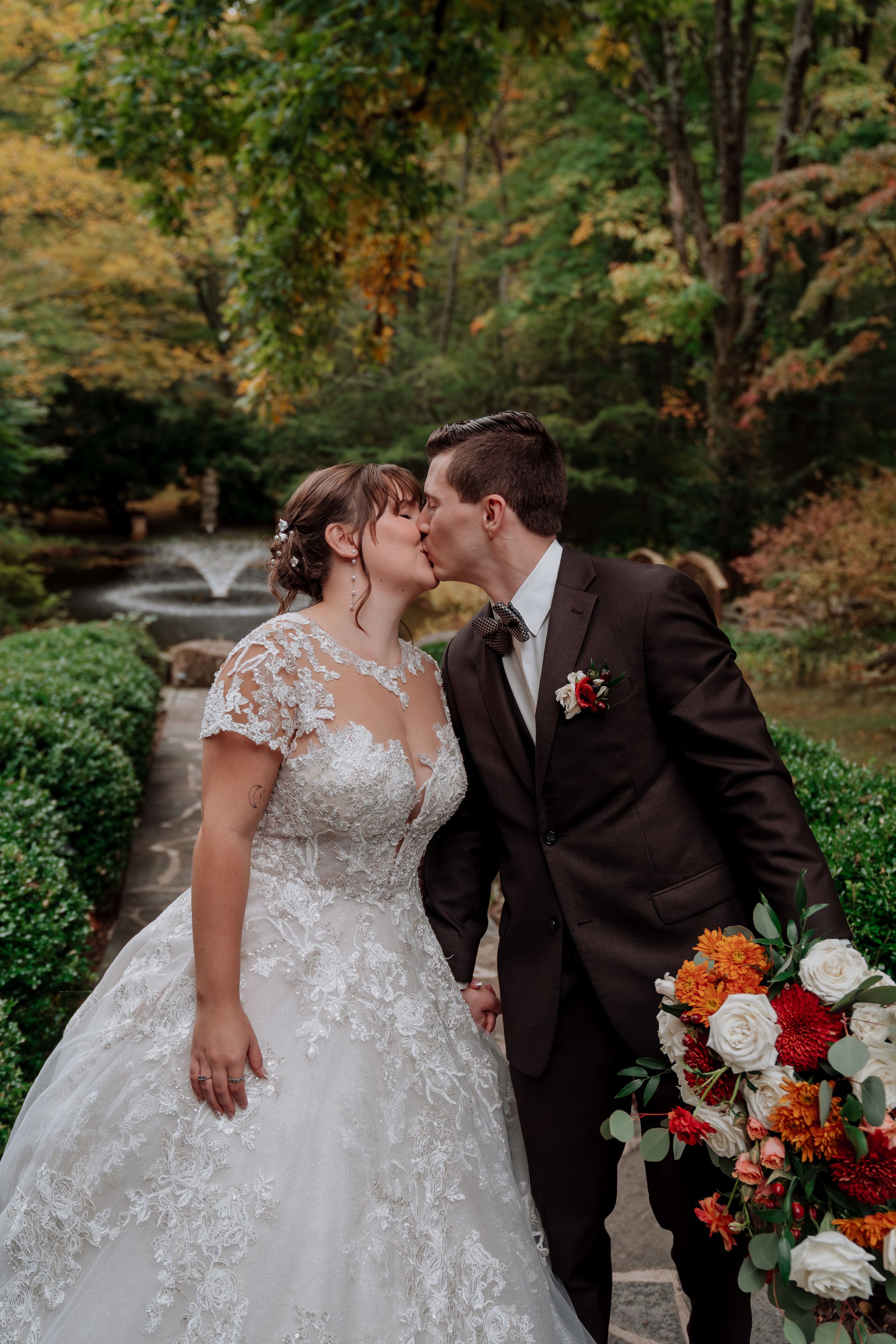 Bride and groom kiss in garden with autumn foliage and fountain backdrop holding fall bouquet.