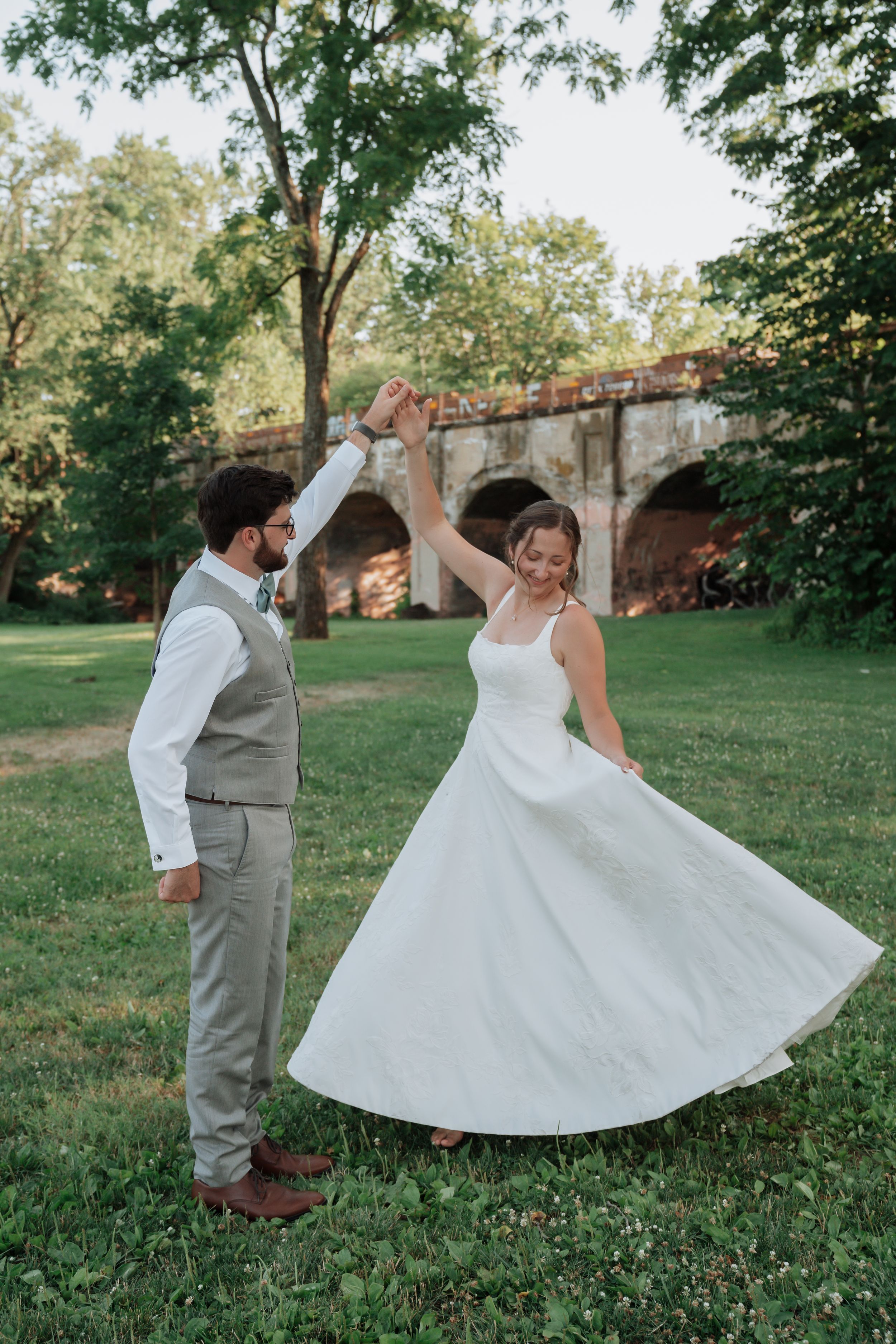 Groom twirling bride in flowing white dress outdoors near a stone bridge in golden evening light.
