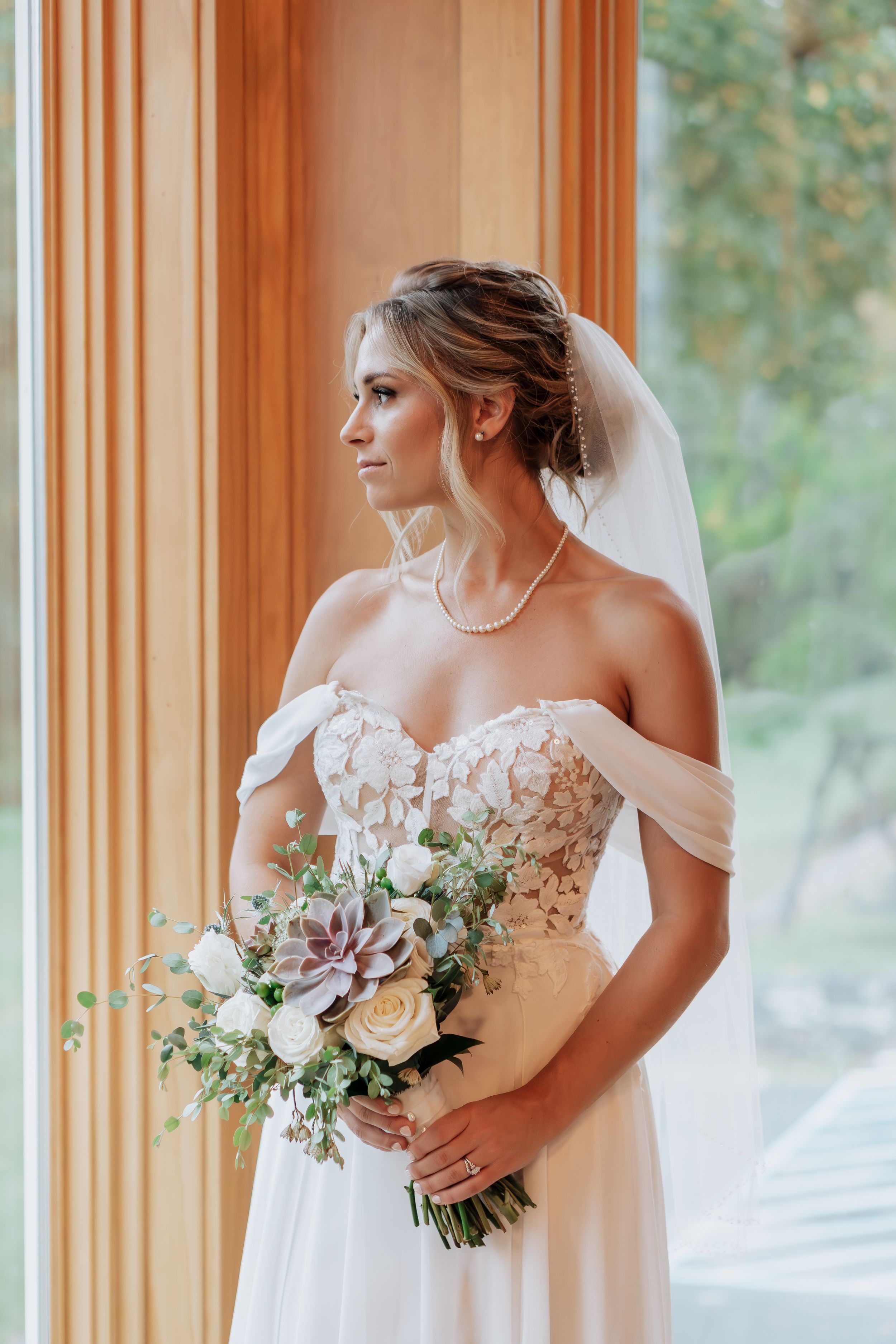 Elegant bride in off-shoulder lace gown holds a bouquet with succulents while gazing out a window.