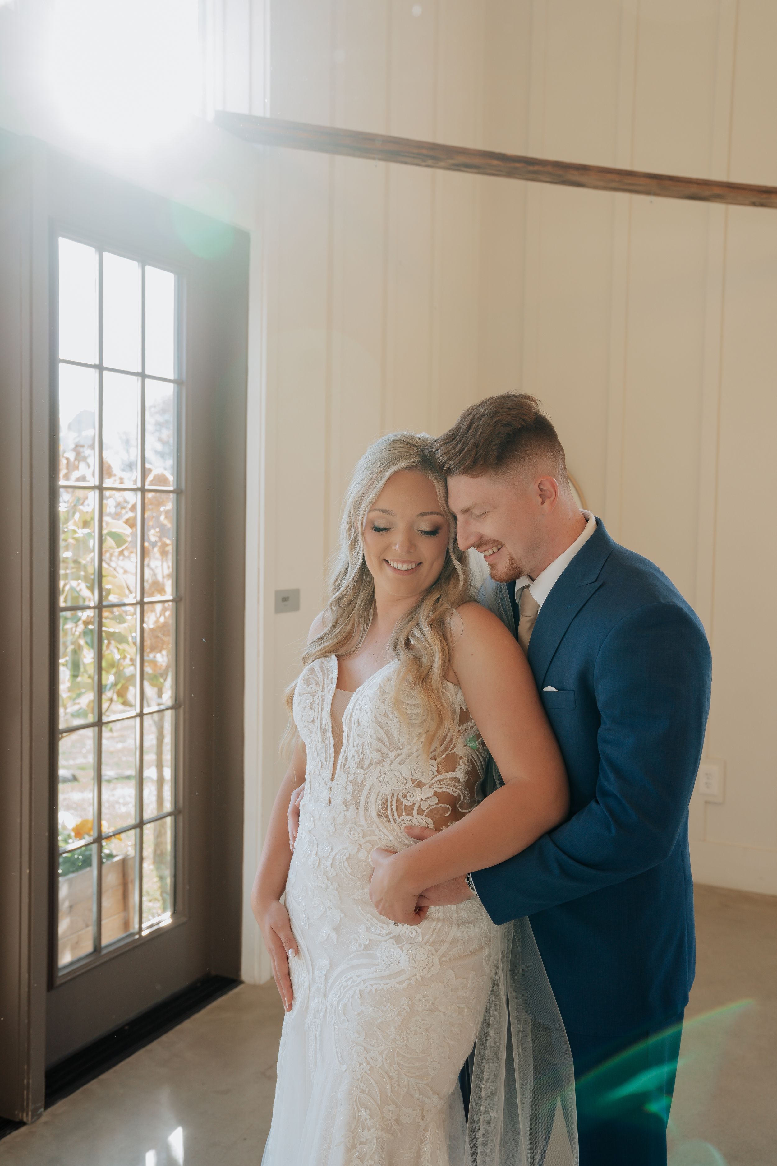 Bride and groom share an intimate embrace near a sunlit door inside a bright white wedding venue.