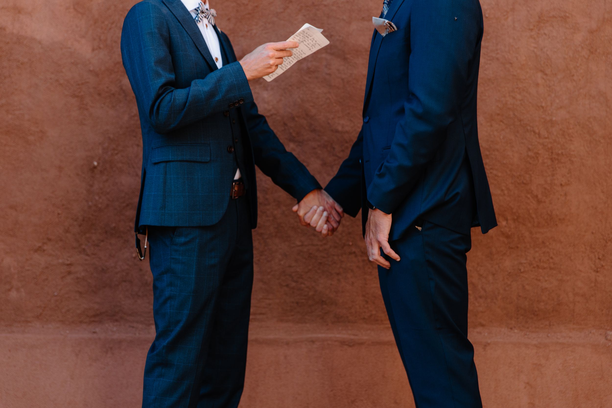 Two people in navy blue suits exchanging vows or reading letters against a terracotta wall.