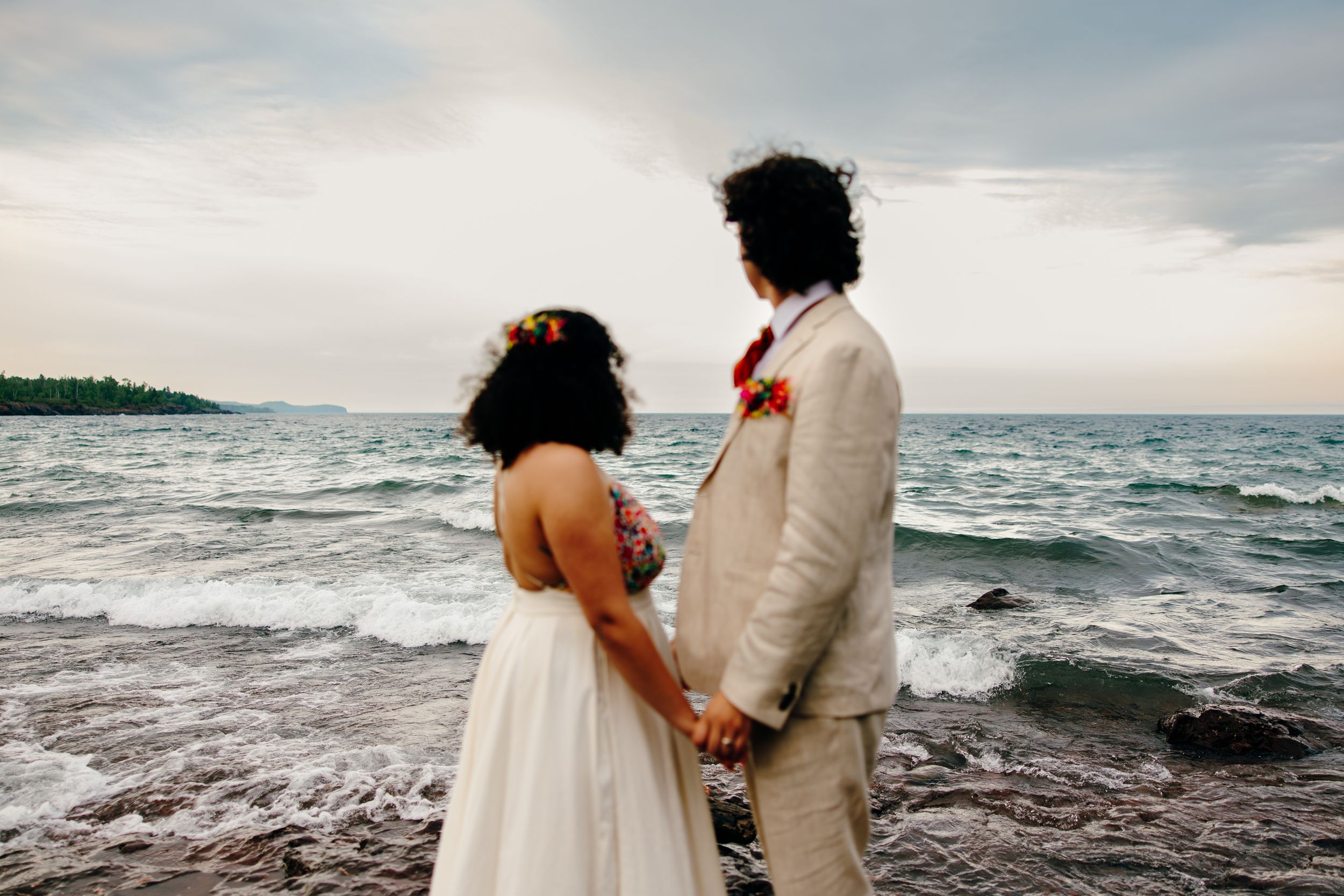 Couple in wedding attire walks along rocky beach shoreline with waves crashing behind them at sunset.