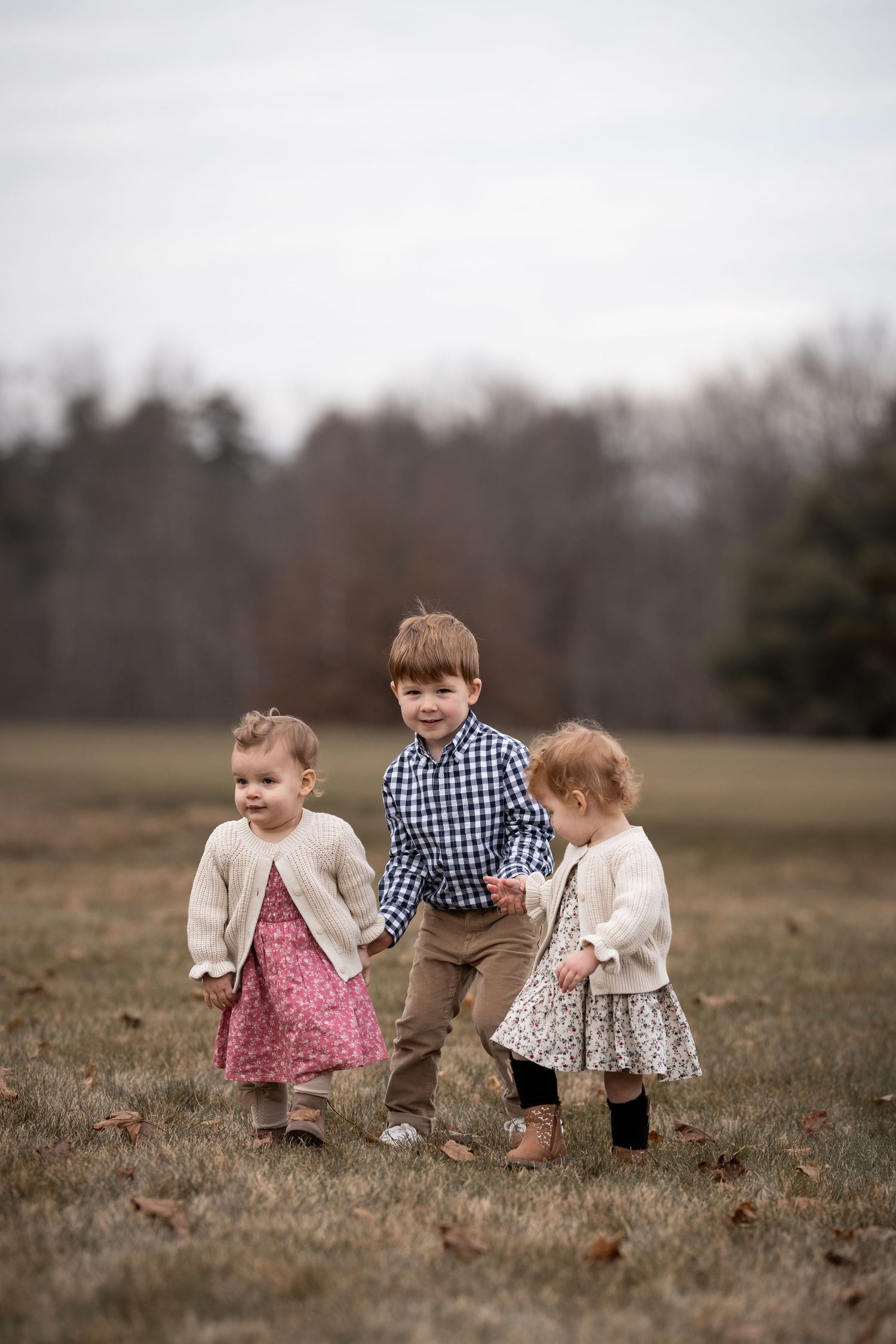 Three children wearing vintage-inspired clothing stand together in an autumn field on an overcast day.
