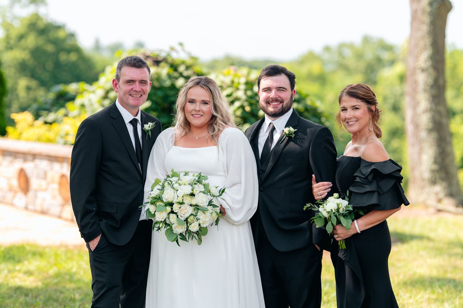 Wedding party of four posing outdoors, bride in white gown with bouquet, groom and parents in elegant black attire.