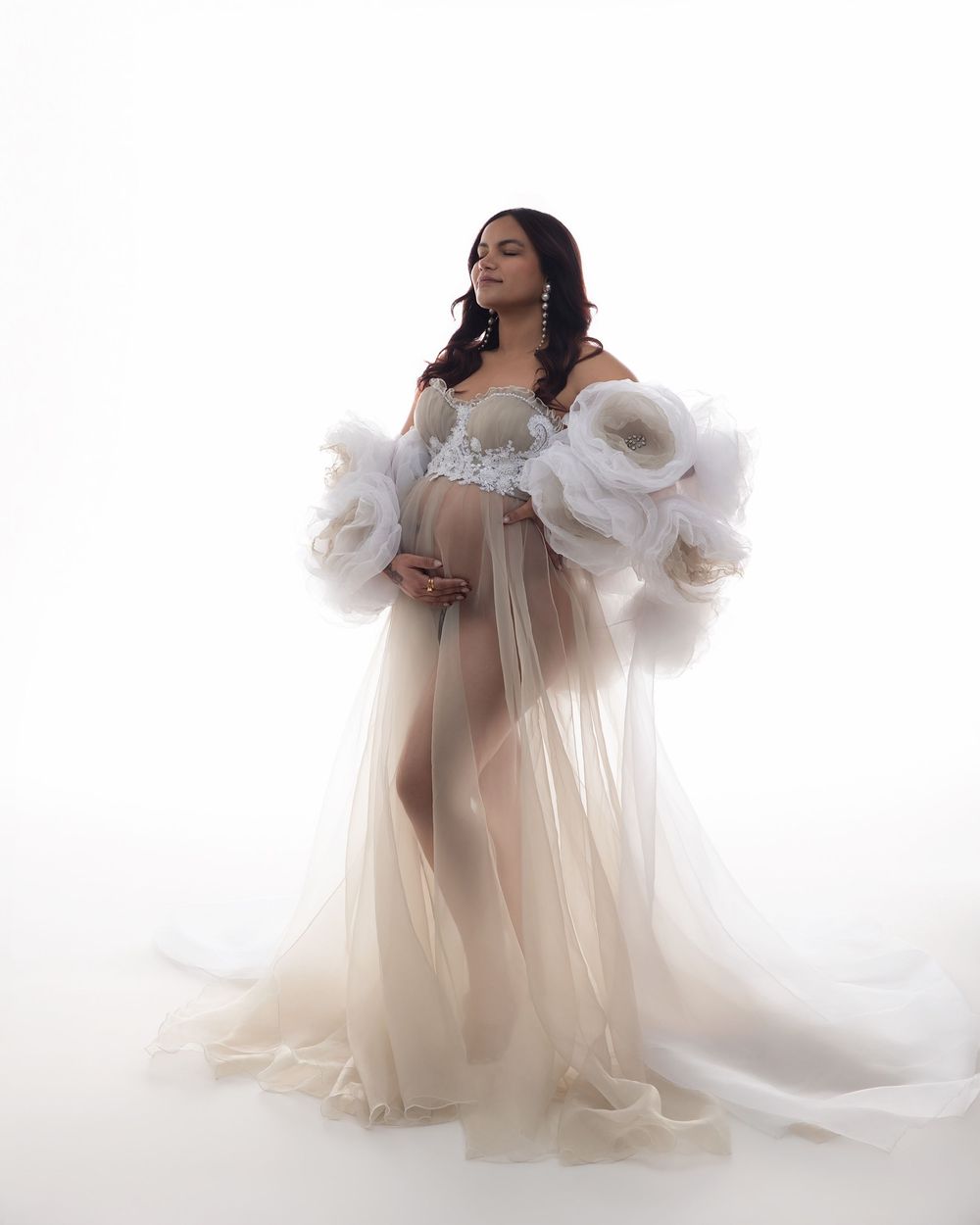 Pregnant woman in elegant white tulle maternity gown with floral details poses gracefully against white background.
