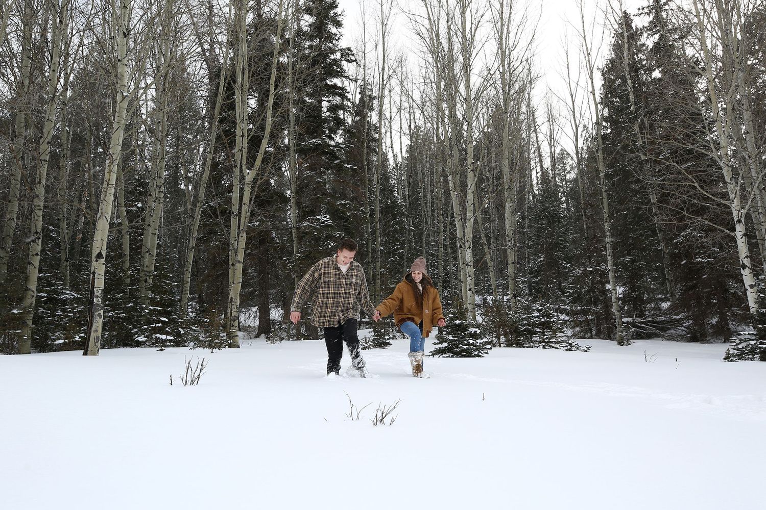 Couple in warm winter jackets holds hands and walks through deep snow among tall evergreen and bare aspen trees in a quiet forest.