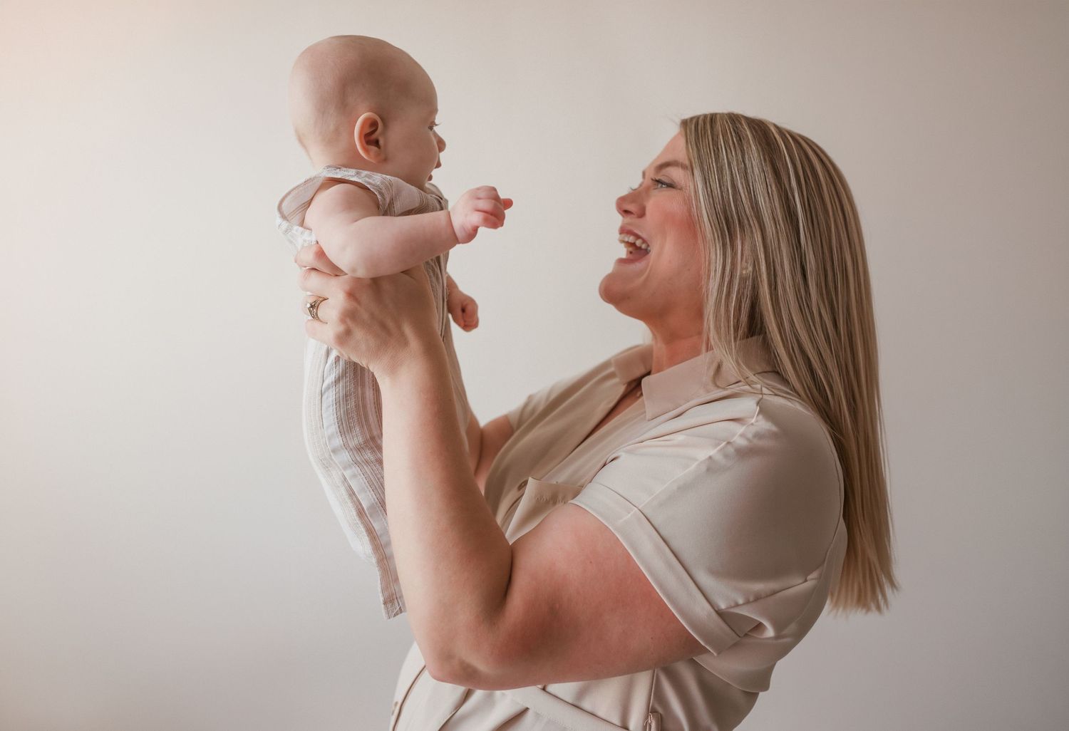 A joyful mother lifts her smiling baby overhead against a light background, sharing a playful bonding moment.